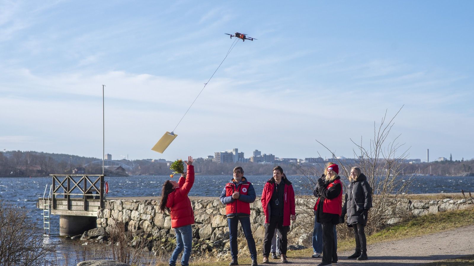En grupp människor står vid vattnet. Magdalena Forsberg tar emot ett guldkuvert som komemr flygande med en drönare.